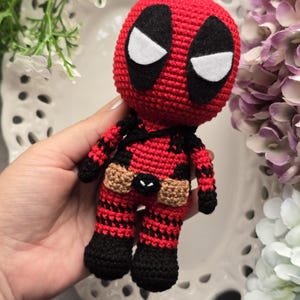 Puede incluir: Un juguete de Deadpool tejido a crochet con un traje rojo y negro, un cinturón marrón y ojos blancos.