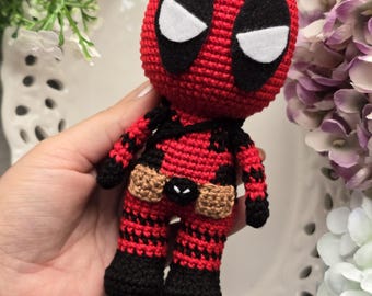 Op de afbeelding: Een gehaakt Deadpool-speelgoedfiguur met een rood-zwart pak, een bruine riem en witte ogen.