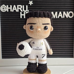 Puede incluir: Figura de amigurumi de crochet de un jugador de fútbol que lleva una camiseta blanca con detalles negros, pantalones cortos negros y botas de fútbol blancas y negras. La camiseta tiene el texto "Fly Emirates" y el número "7". El jugador está sosteniendo un balón de fútbol blanco y negro y está de pie sobre una base de madera.