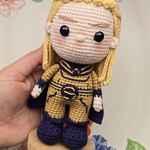 May include: A crocheted doll with fair skin, blonde hair, and a gold and navy blue costume. The doll has large black eyes and pink cheeks. It is mounted on a small wooden base.