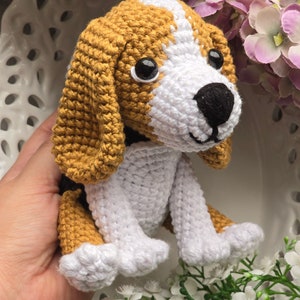 Puede incluir: Un juguete de cachorro beagle de crochet con pelaje marrón y blanco, ojos negros y nariz negra. El juguete está sentado sobre una superficie blanca con flores blancas.