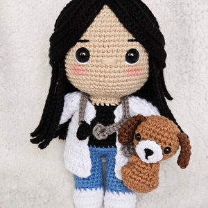 Puede incluir: Muñeca de crochet con una bata blanca, pantalones vaqueros azules y un estetoscopio. La muñeca sostiene un cachorro de crochet marrón.