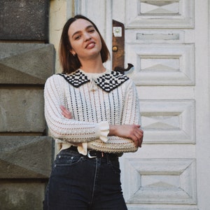 May include: A woman wearing a cream-colored sweater with a black and white checkered collar and dark wash jeans. The sweater has a textured knit design and long sleeves. The jeans are high-waisted with a black belt. The background features a white door and stone wall.