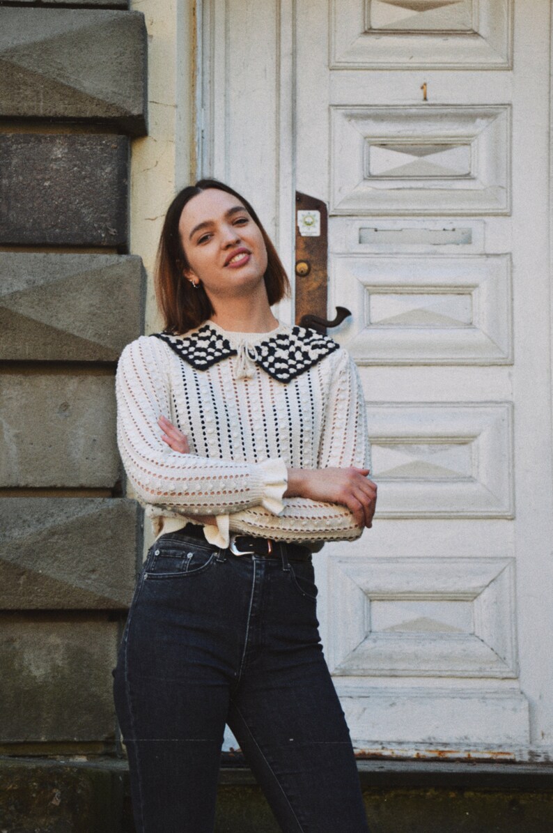 May include: A woman wearing a cream-colored sweater with a black and white checkered collar and dark wash jeans. The sweater has a textured knit design and long sleeves. The jeans are high-waisted with a black belt. The background features a white door and stone wall.