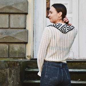 May include: A cream-colored, open-knit sweater with a black and white patterned collar is paired with dark wash denim jeans. The sweater has long sleeves and a textured design. The person is standing in front of a white door.