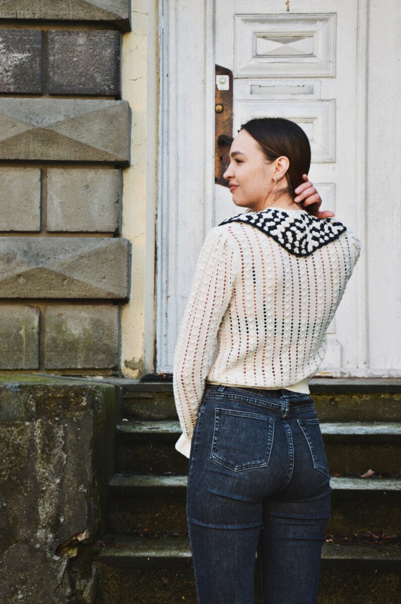 May include: A cream-colored, open-knit sweater with a black and white patterned collar is paired with dark wash denim jeans. The sweater has long sleeves and a textured design. The person is standing in front of a white door.