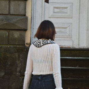 May include: A person wearing a cream-colored, open-weave sweater with a black and white patterned collar and cuffs. The person is also wearing dark blue jeans. The background includes a white door and stone steps.
