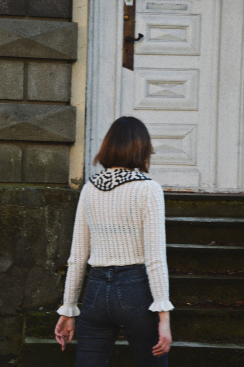 May include: A person wearing a cream-colored, open-weave sweater with a black and white patterned collar and cuffs. The person is also wearing dark blue jeans. The background includes a white door and stone steps.