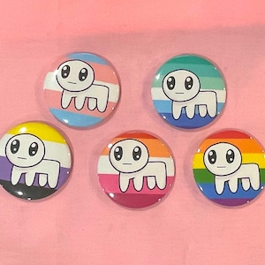 May include: Five round buttons with a white cartoon creature design. Each button features a different LGBTQ+ pride flag design, including transgender, gay, non-binary, lesbian, and rainbow pride flags. The buttons are on a pink background.
