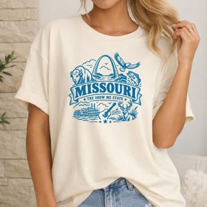 以下が含まれることがあります： クリーム色の半袖Tシャツで、青いグラフィックプリントが施されています。デザインには「MISSOURI THE SHOW ME STATE」という文字、バイソン、ゲートウェイアーチ、ワシ、魚、外輪船、アコースティックギター、シカが描かれています。