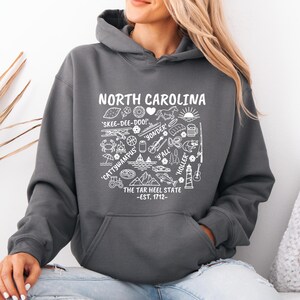 Gildan hooded sweatshirt featuring North Carolina graphic filled with state icons including lighthouse, mountains, fishing rod, crab, tractor, sailboat, guitar, sunrise, and regional phrases. Front design displayed on seated female model wearing ripped denim against ocean background. Cozy Tar Heel hoodie created for coastal mountain lifestyle, regional pride, souvenir memories, and casual daily wear.