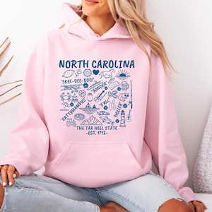 Gildan hooded sweatshirt featuring North Carolina graphic filled with state icons including lighthouse, mountains, fishing rod, crab, tractor, sailboat, guitar, sunrise, and regional phrases. Front design displayed on seated female model wearing ripped denim against ocean background. Cozy Tar Heel hoodie created for coastal mountain lifestyle, regional pride, souvenir memories, and casual daily wear.
