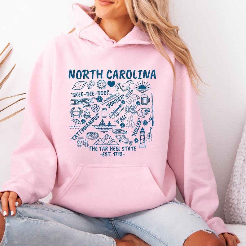 Gildan hooded sweatshirt featuring North Carolina graphic filled with state icons including lighthouse, mountains, fishing rod, crab, tractor, sailboat, guitar, sunrise, and regional phrases. Front design displayed on seated female model wearing ripped denim against ocean background. Cozy Tar Heel hoodie created for coastal mountain lifestyle, regional pride, souvenir memories, and casual daily wear.