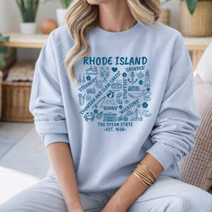 Könnte beinhalten: Hellblaues Sweatshirt mit einem grafischen Design. Das Design enthält den Text "RHODE ISLAND" und Illustrationen lokaler Symbole, wie einen Hummer, Muschelkuchen und ein Segelboot. Der Text "The Ocean State - EST. 1636-" ist ebenfalls enthalten.