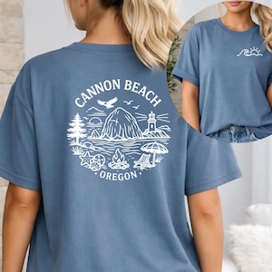 Peut inclure: T-shirt bleu poussiéreux avec un motif blanc au dos. Le motif représente une scène côtière avec un phare, des oiseaux et une formation rocheuse, avec l'inscription "Cannon Beach Oregon". Un petit motif de vagues et de soleil est sur le devant.