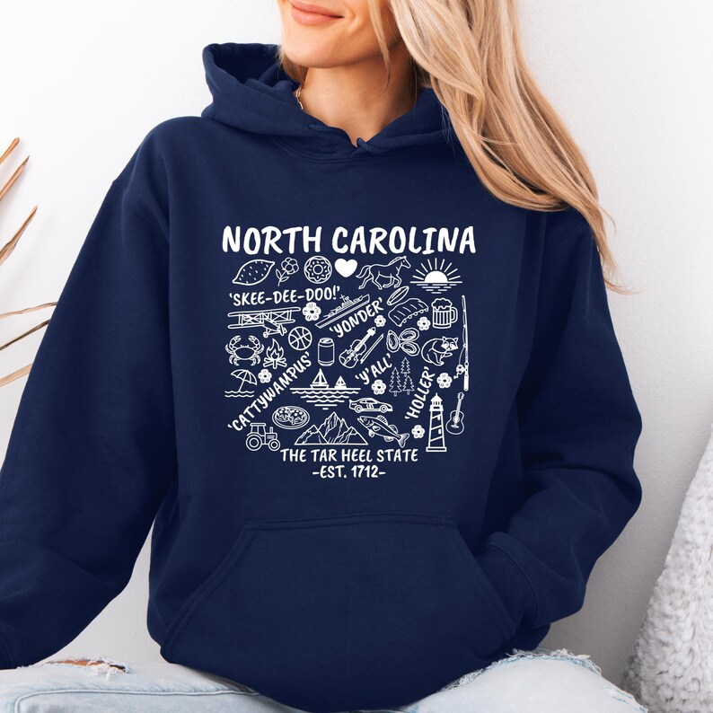 Gildan hooded sweatshirt featuring North Carolina graphic filled with state icons including lighthouse, mountains, fishing rod, crab, tractor, sailboat, guitar, sunrise, and regional phrases. Front design displayed on seated female model wearing ripped denim against ocean background. Cozy Tar Heel hoodie created for coastal mountain lifestyle, regional pride, souvenir memories, and casual daily wear.