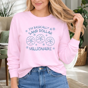May include: A light pink long-sleeved shirt with the text "I'm Basically a Sand Dollar Millionaire" in teal. The design includes three sand dollar illustrations. The shirt is paired with blue jeans.