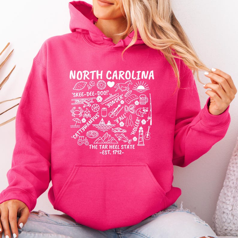 Gildan hooded sweatshirt featuring North Carolina graphic filled with state icons including lighthouse, mountains, fishing rod, crab, tractor, sailboat, guitar, sunrise, and regional phrases. Front design displayed on seated female model wearing ripped denim against ocean background. Cozy Tar Heel hoodie created for coastal mountain lifestyle, regional pride, souvenir memories, and casual daily wear.