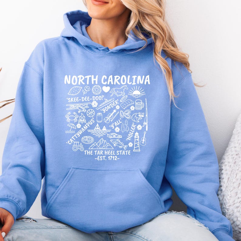 Gildan hooded sweatshirt featuring North Carolina graphic filled with state icons including lighthouse, mountains, fishing rod, crab, tractor, sailboat, guitar, sunrise, and regional phrases. Front design displayed on seated female model wearing ripped denim against ocean background. Cozy Tar Heel hoodie created for coastal mountain lifestyle, regional pride, souvenir memories, and casual daily wear.