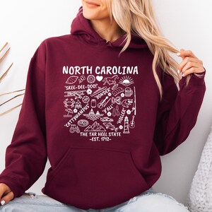 Gildan hooded sweatshirt featuring North Carolina graphic filled with state icons including lighthouse, mountains, fishing rod, crab, tractor, sailboat, guitar, sunrise, and regional phrases. Front design displayed on seated female model wearing ripped denim against ocean background. Cozy Tar Heel hoodie created for coastal mountain lifestyle, regional pride, souvenir memories, and casual daily wear.