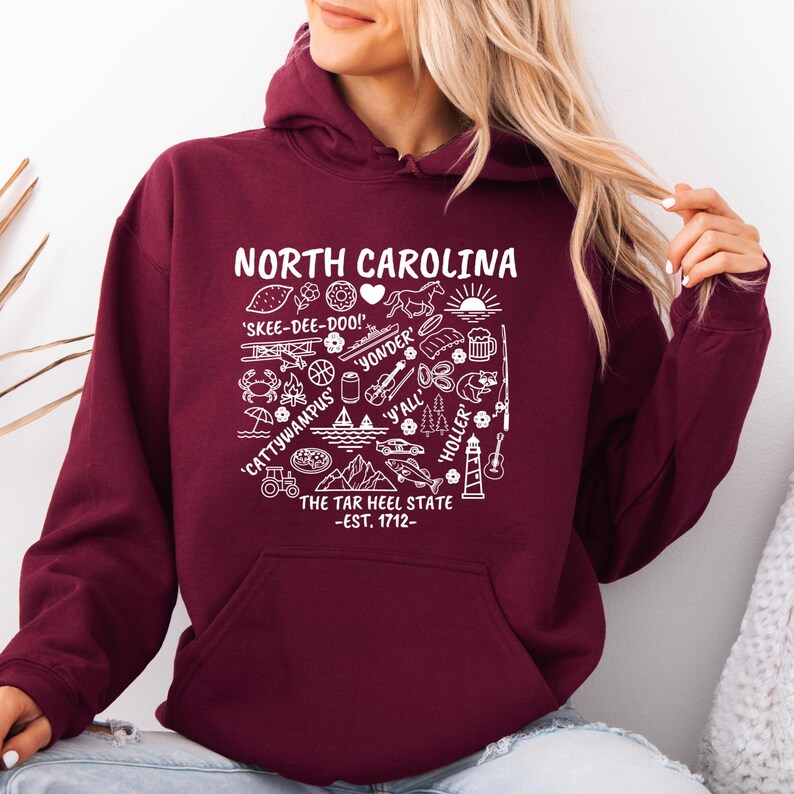 Gildan hooded sweatshirt featuring North Carolina graphic filled with state icons including lighthouse, mountains, fishing rod, crab, tractor, sailboat, guitar, sunrise, and regional phrases. Front design displayed on seated female model wearing ripped denim against ocean background. Cozy Tar Heel hoodie created for coastal mountain lifestyle, regional pride, souvenir memories, and casual daily wear.