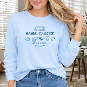 May include: Light blue long-sleeve shirt with the text "PROFESSIONAL SEASHELL COLLECTOR HIGHLY SKILLED" in teal. The design includes seashell illustrations. The shirt is made of a soft material.