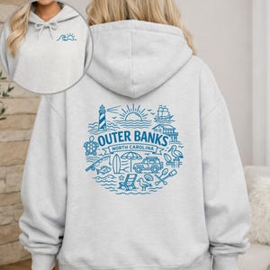 Könnte beinhalten: Ein hellgrauer Kapuzenpullover mit einer blauen Grafik auf der Rückseite und dem Schriftzug Outer Banks North Carolina, umgeben von Illustrationen eines Leuchtturms, einer Sonne, eines Schiffes, einer Schildkröte und eines Fahrzeugs.
