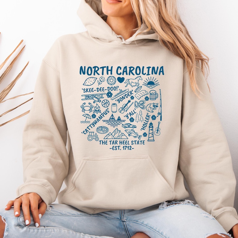 Gildan hooded sweatshirt featuring North Carolina graphic filled with state icons including lighthouse, mountains, fishing rod, crab, tractor, sailboat, guitar, sunrise, and regional phrases. Front design displayed on seated female model wearing ripped denim against ocean background. Cozy Tar Heel hoodie created for coastal mountain lifestyle, regional pride, souvenir memories, and casual daily wear.