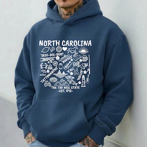 Gildan hooded sweatshirt featuring North Carolina graphic filled with state icons including lighthouse, mountains, fishing rod, crab, tractor, sailboat, guitar, sunrise, and regional phrases. Front design displayed on seated female model wearing ripped denim against ocean background. Cozy Tar Heel hoodie created for coastal mountain lifestyle, regional pride, souvenir memories, and casual daily wear.