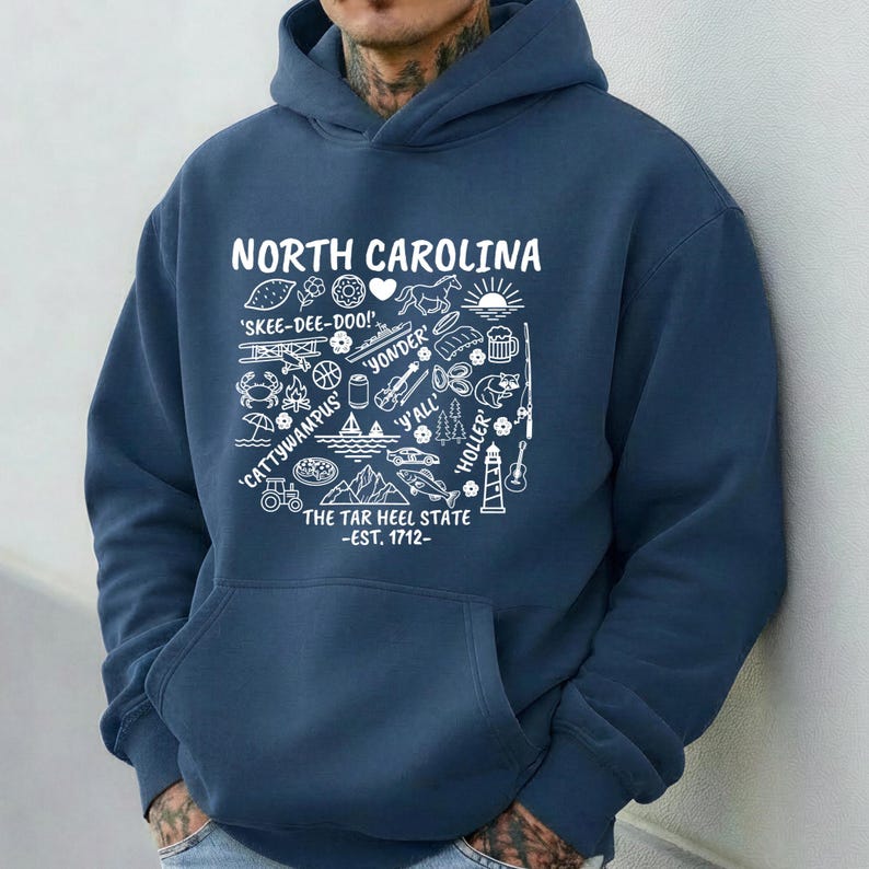 Gildan hooded sweatshirt featuring North Carolina graphic filled with state icons including lighthouse, mountains, fishing rod, crab, tractor, sailboat, guitar, sunrise, and regional phrases. Front design displayed on seated female model wearing ripped denim against ocean background. Cozy Tar Heel hoodie created for coastal mountain lifestyle, regional pride, souvenir memories, and casual daily wear.