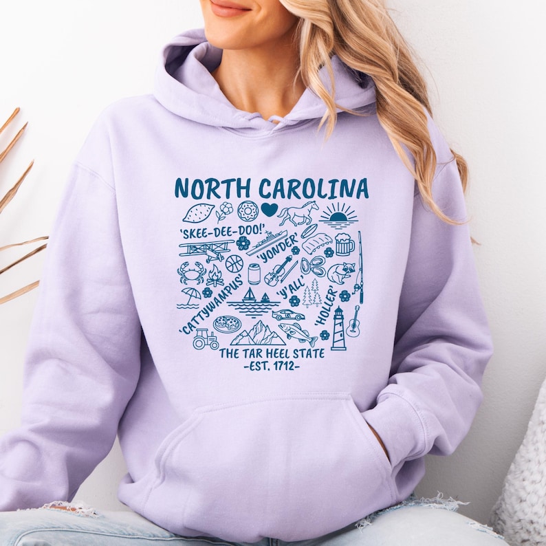 Gildan hooded sweatshirt featuring North Carolina graphic filled with state icons including lighthouse, mountains, fishing rod, crab, tractor, sailboat, guitar, sunrise, and regional phrases. Front design displayed on seated female model wearing ripped denim against ocean background. Cozy Tar Heel hoodie created for coastal mountain lifestyle, regional pride, souvenir memories, and casual daily wear.