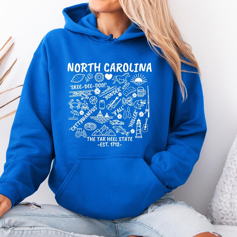 Gildan hooded sweatshirt featuring North Carolina graphic filled with state icons including lighthouse, mountains, fishing rod, crab, tractor, sailboat, guitar, sunrise, and regional phrases. Front design displayed on seated female model wearing ripped denim against ocean background. Cozy Tar Heel hoodie created for coastal mountain lifestyle, regional pride, souvenir memories, and casual daily wear.