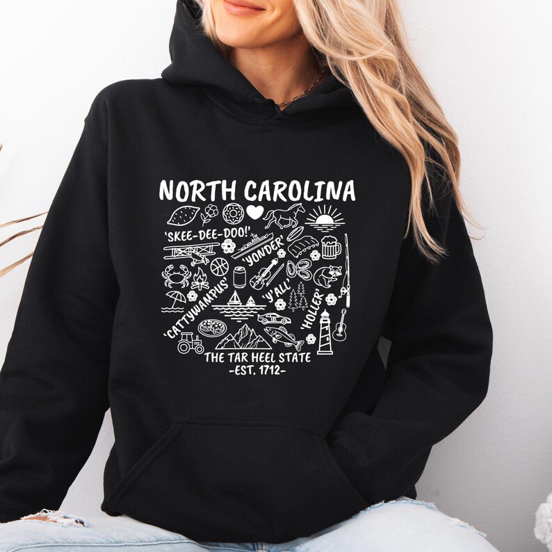Gildan hooded sweatshirt featuring North Carolina graphic filled with state icons including lighthouse, mountains, fishing rod, crab, tractor, sailboat, guitar, sunrise, and regional phrases. Front design displayed on seated female model wearing ripped denim against ocean background. Cozy Tar Heel hoodie created for coastal mountain lifestyle, regional pride, souvenir memories, and casual daily wear.