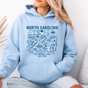 Gildan hooded sweatshirt featuring North Carolina graphic filled with state icons including lighthouse, mountains, fishing rod, crab, tractor, sailboat, guitar, sunrise, and regional phrases. Front design displayed on seated female model wearing ripped denim against ocean background. Cozy Tar Heel hoodie created for coastal mountain lifestyle, regional pride, souvenir memories, and casual daily wear.