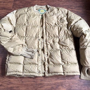 May include: A tan quilted jacket with a round collar and button closure. The jacket has long sleeves with thumb holes at the cuffs. The jacket is made of a lightweight material and is likely designed for outdoor activities.