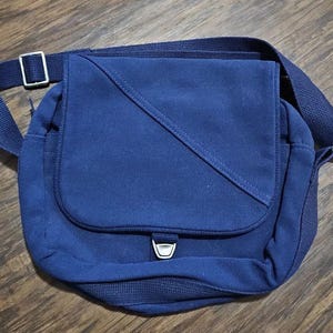 May include: A navy blue messenger bag with a flap closure and adjustable shoulder strap. The bag features a silver-colored buckle and a diagonal stitched design on the front. The bag is made of a sturdy fabric.