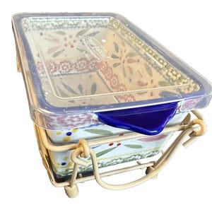 May include: Rectangular ceramic baking dish with a floral pattern, a clear plastic lid, and a blue locking mechanism. The dish is set in a cream-colored metal stand. The dish has a floral pattern with red, green, and yellow accents.