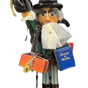 May include: A wooden nutcracker figurine of a man in a black top hat and green coat, holding a small blue book that says "Scrooge & Marley". The figurine has a stern expression and carries a locked orange chest.
