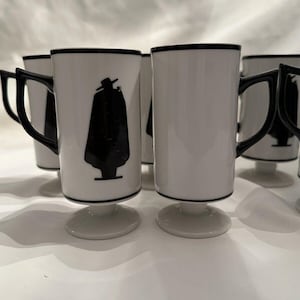 May include: Set of four white ceramic mugs with black trim and a black silhouette of a person wearing a hat and a cape. The mugs have a pedestal base.