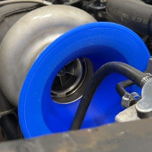 May include: Close-up of an engine component featuring a silver-colored turbocharger with a bright blue intake. Black hoses and various engine parts surround the turbocharger, creating a detailed view of the engine's mechanics.