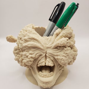 Puede incluir: Un organizador de escritorio impreso en 3D de color beige con forma de cabeza de monstruo gritando con la boca abierta y dientes visibles. Dos bol&iacute;grafos, uno negro y otro verde, se insertan en la boca.
