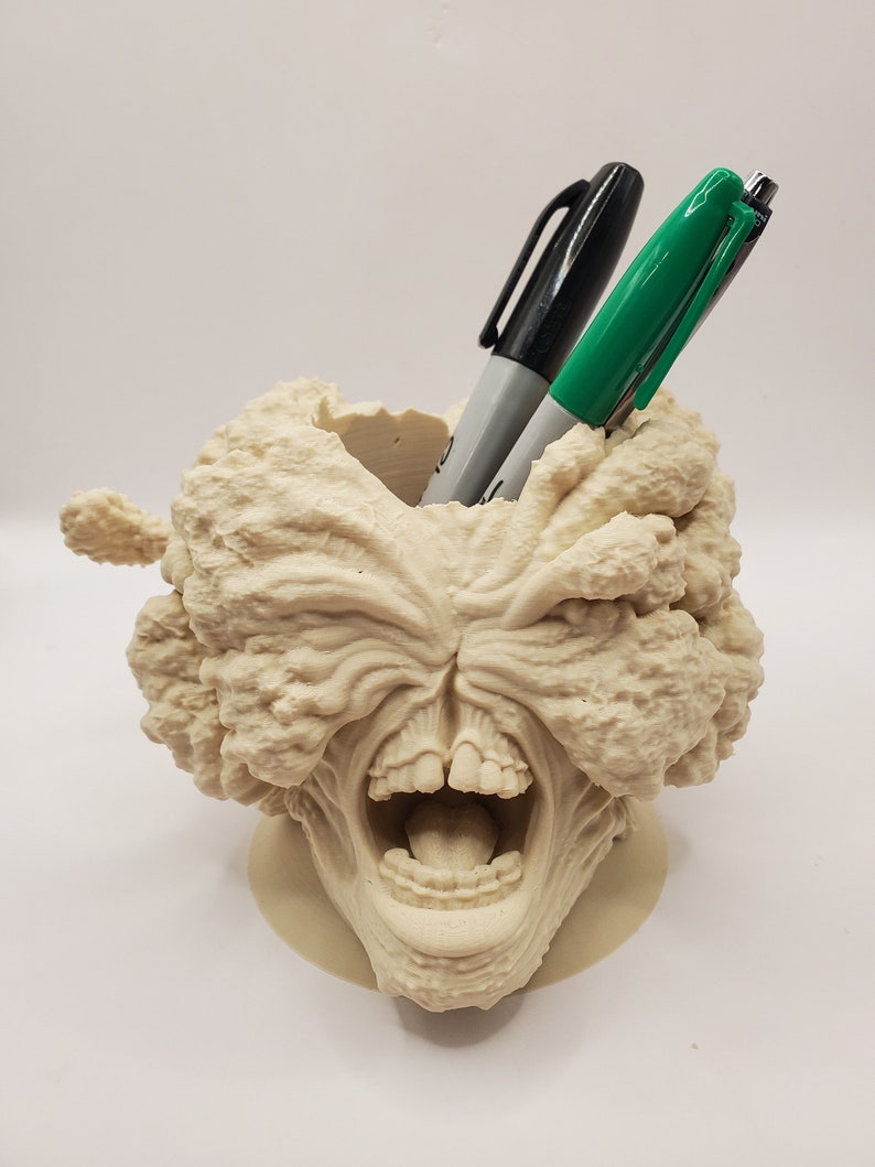 Puede incluir: Un organizador de escritorio impreso en 3D de color beige con forma de cabeza de monstruo gritando con la boca abierta y dientes visibles. Dos bol&iacute;grafos, uno negro y otro verde, se insertan en la boca.