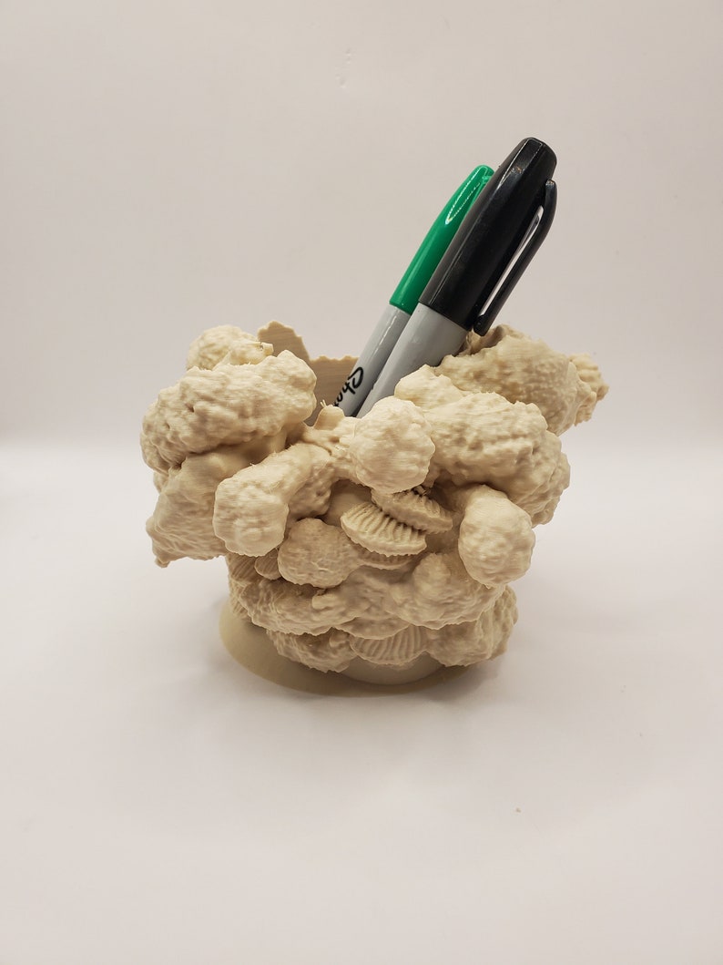 Puede incluir: Un organizador de escritorio de color beige en forma de concha impreso en 3D con dos marcadores negros y verdes en su interior.
