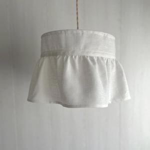 May include: A white fabric lampshade with a ruffled skirt design, suspended from a natural-colored cord. The lampshade has a cylindrical top and a flared, gathered bottom, creating a soft, textured appearance. The light fixture is against a plain white background.
