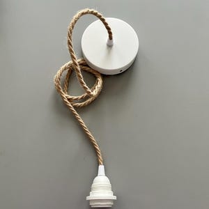 Puede incluir: Una lámpara de techo blanca con un cable de cuerda natural. La lámpara incluye una base circular blanca y un portalámparas blanco. El cable de cuerda es de color marrón claro y está enrollado.