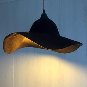 May include: A black pendant light fixture with a wide, hat-like shade. The interior of the shade has a warm, golden hue, contrasting with the dark exterior. The light hangs from a thin, off-white cord.