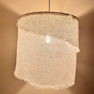 Pu&ograve; includere: Un paralume per lampada in tessuto bianco con un bordo sfrangiato appeso a un cavo. Il paralume &egrave; cilindrico e ha una lampadina all'interno.