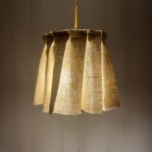 May include: A pendant light fitting featuring a cylindrical lampshade made of light tan, textured hessian fabric with vertical pleats and a frayed bottom edge, suspended from a single, twisted, bright yellow electrical cable.