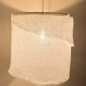 Pu&ograve; includere: Un paralume per lampada in tessuto bianco con un bordo sfrangiato appeso a un cavo. Il paralume &egrave; acceso ed emana una luce calda.