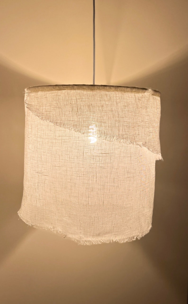 Pu&ograve; includere: Un paralume per lampada in tessuto bianco con un bordo sfrangiato appeso a un cavo. Il paralume &egrave; acceso ed emana una luce calda.